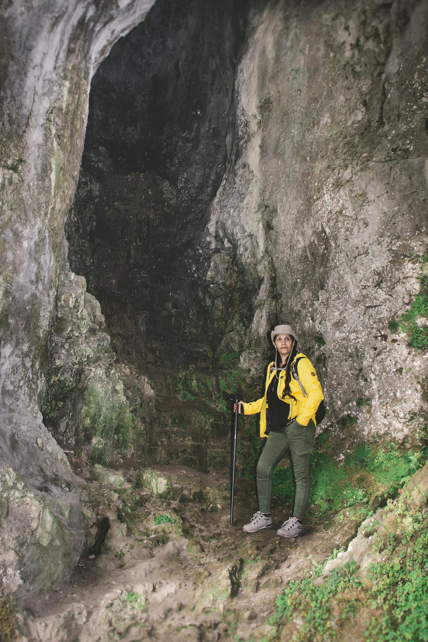 A woman in a yellow jacket explores a rocky cave with a hiking cane, enjoying nature.